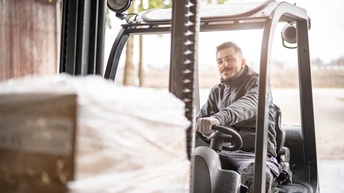 Eine Person in einer grauen Weste, mit kurzen, dunklen Haaren und einem Bart lenkt einen Gabelstapler und blickt direkt in die Kamera. Vorne am Gabelstapler sind Kisten