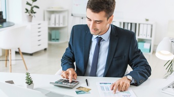 Eine Person im Anzug sitzt an einem weißen Tisch. Vor sich hat er einen Taschenrechner, einen Stift, einen aufgeklappten Laptop und Papiere mit Statistiken. In dem Raum ist noch ein Schreibtisch, Regale mit Ordnern