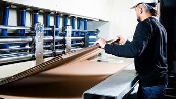Eine Person steht an einer großen Industriemaschine und hält mit beiden Händen einen breiten Stapel Wellpappe. Sie trägt einen Schutzhelm und eine Schutzbrille. Daneben ist eine geöffnete Maschine mit mehreren waagrechten, silberfarbenen Metallwalzen