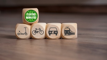 Vier Holwürfel mit Symbolen E-Roller, Fahrrad, Elektroauto und Zug nebeneinander platziert, oben auf ein weiterer Holzwürfel mit weißem Schriftzug Elektromobilität in grünem Kreis