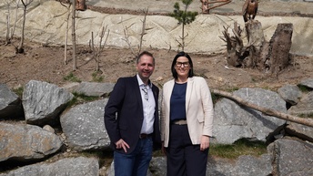 Zwei Erwachsene stehen vor einem großen Gehege mit mehreren Geiern, die auf Holzstämmen sitzen. Die Personen sind gepflegt gekleidet und lächeln in die Kamera. Im Hintergrund sind Felsen, Bäume und ein abgesperrter Naturbereich zu sehen.
