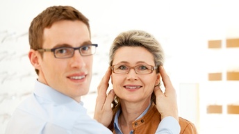 Ein Optiker setzt einer Kundin eine neue Brille zum Probieren auf, beide blicken dabei lächelnd in die Kamera