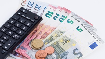 Eine Fernbedienung liegt auf nebeneinander aufgelegten Geldscheinen am Tisch, daneben sind noch ein paar Münzen gestapelt.