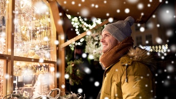 Person mit grauer Haube, Bart, gelber Jacke und orangem Schal blickt freudig auf eine Auslage eines Standes, der hell erleuchtet mit Christbaumschmuck behangen ist, ringsum sind Schneeflocken