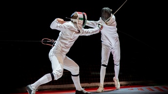 Zwei Personen in Fechtkleidung und mit Fechthelmen mit erhobenen Degen und in Bewegung