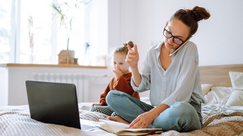 Eine Person mit dunklen, geschlossenen Haaren und Brille sitzt in einem Bett und telefoniert. Vor ihr liegt ein Notizbuch. Neben ihr sitzt ein Kind, das auf den Bildschirm eines aufgeklappten Laptops blickt