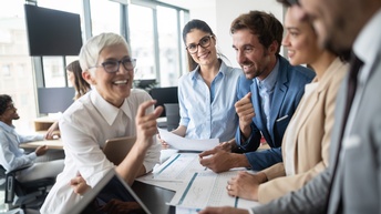Personen in Businesskleidung stehen freudig rund um einen Stehtisch während Unterlagen sowie Laptop und Notebook im Einsatz sind