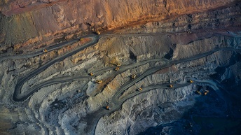 Aufnahme eines Bergwerks aus der Luft mit verschlungenen Wegen, die vom Rand des Werks nach unten führen. Auf den Wegen fahren mehrere Fahrzeuge