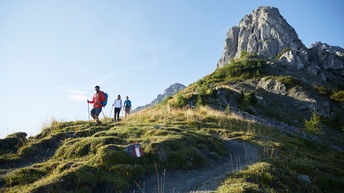 Drei Personen in Wanderkleidung, zwei davon mit Wanderstöcken ausgerüstet, marschieren Berg herunter