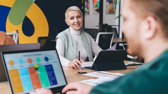 Person mit kurzen blonden Haaren sitzt an einem Tisch mit Tablet und Unterlagen und blickt auf eine Person ihr gegenüber in der Unschärfe, weitere Personen sitzen am Tisch, im Hintergrund zeigt sich ein modernes Büro