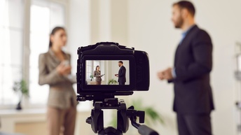 Im Hintergrund stehen sich zwei Personen in Sakkos und Stoffhose gegenüber, die sich miteinander unterhalten. Im Vordergrund ist eine Kamera auf einem Stativ. Am Bildschirm der Kamera sind die zwei Personen