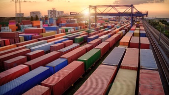 Aufsicht auf einen großen Containerumschlagplatz bei Sonnenuntergang. In der Mitte befindet sich ein großer Kran. Rechts daneben sind Schienen zu sehen. Links, weit im Hintergrund, befinden sich einige Wohngebäude.