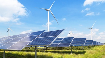 Mehrere Reihen von Solarpanelen auf Metallgerüsten auf einem Feld, dahinter Windräder unter blauem Himmel mit vereinzelten Wolken