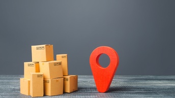 Miniaturen von Kartonagenschachteln aufeinander gestapelt, daneben rotes Standortsymbol aus Holz vor dunklem Hintergrund