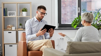 Rückenansicht einer Person mit kurzen, grauen Haaren, die auf einem Sofa sitzt. Ihr gegenüber sitzt eine andere Person mit Bart, kurzen, braunen Haaren und Brille, die auf ein Tablet blickt, das sie hält