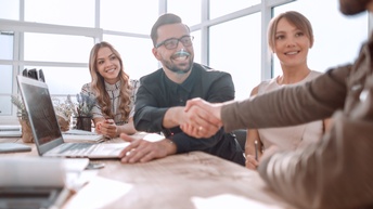 Mehrere lächelnde Personen sitzen an Tisch mit Laptop und Dokumenten, zwei Personen reichen sich die Hände