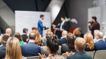Rückenansicht eines Publikums in mehreren Sesselreihen, die klatschen. Ihnen gegenüber stehen sich zwei Personen vor einem Banner gegenüber. Rechts neben dem Publikum steht eine Person mit Kamera