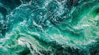 Luftaufnahme von turbulenten, schäumenden Wellen im Meer. Die Farben reichen von tiefem Blau bis zu hellem Türkis, und die Wellen bilden komplexe Muster und Strudel