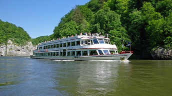 Auf einem Fluss fährt ein zweistöckiges, langes Boot. Am obersten Deck des Bootes stehen Personen. Links und rechts vom Fluss sind Steine sowie grüne Bäume. Der Himmel ist blau