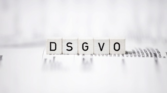 Weiße Würfel mit dem Aufdruck DSGVO, die auf einem textlichen Papier aufliegen und ringsum zeigt sich Unschärfe