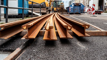 Im Vordergrund liegen mehrere gebogene Stahlschienen parallel auf Holzunterlagen auf einer asphaltierten Straße. Die Schienen sind rostfarben. Im Hintergrund befinden sich Baumaschinen, darunter ein Bagger, sowie Container und Absperrelemente