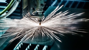Nahaufnahme einer CNC-Lasermaschine, die eine Metallplatte schneidet. Dabei entstehen Funken und grelle Strahlen