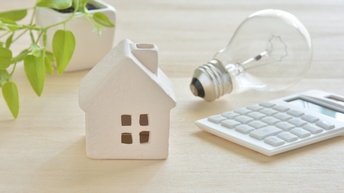 Weißes Keramikhäuschen mit Glühbirne, Taschenrechner und Zierpflanze auf Holztisch