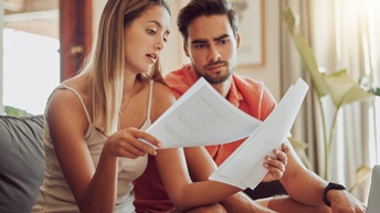 Zwei Personen sitzen auf einer Couch, eine Person hält Unterlagen in der Hand und beide blicken darauf