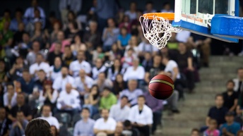 Detailansicht eines Balles der durch Basketballkorb gefallen ist, im Hintergrund verschwommen Publikumstribünen mit Menschen