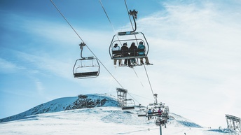 Auf einem Sessellift sitzen auf mehreren Sesseln sitzen Personen in Skianzügen und mit Ski an den Füßen. Unter ihnen sind schneebedeckte Pisten, auf denen vereinzelte Personen sind. Im Hintergrund sieht man schneebedeckte Berge und blauen Himmel.