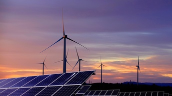 Im Vordergrund sind Solaranlagen auf einer Wiese. Dahinter sind Windräder im Licht der Dämmerung.