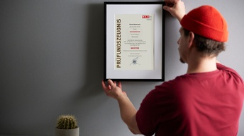 Person mit kurzen dunklen Haaren, Bart, roter Mütze und rotem Shirt steht vor einer grauen Wand und hält einen Bilderrahmen mit Passepartout und Prüfungszeugnis Meister in Händen, daneben steht eine Pflanze