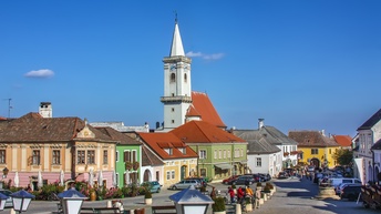 Auf einem asphaltierten Platz stehen ein paar Leute sowie Sitzbänke. Um den Platz stehen alte, niedrige Häuser mit teilweise bunten Fassaden. Zwischen ihnen ragt ein Kirchturm in die Höhe. Der Himmel ist blau