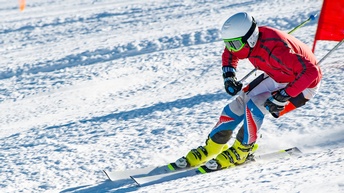Eine Person auf Skiern im Skianzug sowie mit Skihelm und Skibrelle fährt eine Schneepiste hinunter
