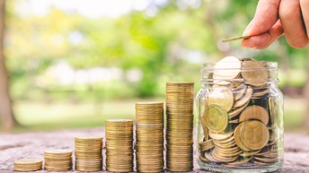 Sechs immer höher werdende Münzstapel stehen auf grauem Untergrund. Neben dem letzten Stapel steht ein großes, gläsernes Gefäß, befüllt mit Münzen, über dem die Hand einer Person eine Münze hält.