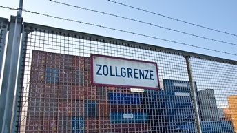 Auf einem Zaun ist ein Schild befestigt, auf dem Zollgrenze steht. Über dem Zaun ist ein Maschendrahtzaun. Dahinter stehen mehrere gestapelte Container