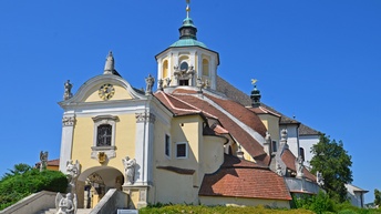 Auf einem leichten Hügel steht eine hellgelbe Kirche mit einem niedrigen Kirchturm. Zur Kirche führen Treppen. Vor dem Eingang und auf dem Geländer sind Engelstatuen. Der Himmel ist blau