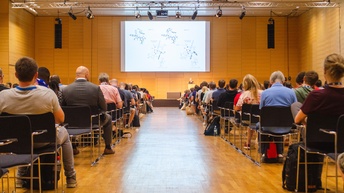 In einem großen Raum sind mehrere Personen, die in vielen Sesselreihen neben- und hintereinander sitzen. Ihnen gegenüber ist eine Person hinter einem Rednerpult. Neben ihr ist eine Leinwand.
