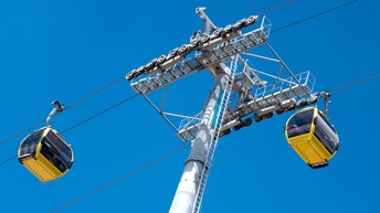 Zwei gelbe Seilbahngondeln unter blauem Himmel an Drahtseil hängend, dazwischen Stahlkonstruktion mit Spulen
