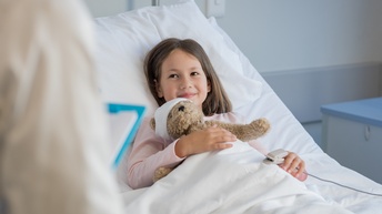 Ein Kind liegt in einem Bett mit weißer Bettwäsche. Auf ihrem Oberkörper liegt ein Teddybär mit verbundenem Kopf. Am linken Finger des Kindes ist ein hellgraues Objekt