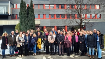 Eine größere Gruppe von Personen steht nebeneinander im Freien vor einem mehrstöckigen Gebäude. Die Personen tragen winterliche Kleidung und blicken in die Kamera. Einige halten Jacken oder Taschen in den Händen, alle stehen dicht beieinander.