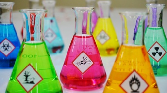 Laborgläser mit verschieden farbenen Flüssigkeiten und unterschiedlichen Aufklebern, mittig im Fokus Laborglas mit pinker Flüssigkeit und weißem quadratischen Aufkleber mit roter Umrandung und schwarzem Symbol für Explosivität: Zerberstende Kugel