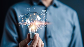 Eine Person in einem blauem Hemd hält eine Glühbirne in die Kamera. Neben der Glühbirne sind Grafiken von Zahnrädern