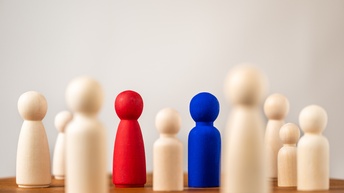 Aufgestellte Spielfiguren aus Holz, von denen 2 rot und blau sowie im Fokus sind. Sie stehen auf einem braunen Holztisch