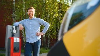 Eine Person steht lachend an eine Ladesäule gestützt, im Vordergrund im Ausschnitt verschwommen ein gelbes Auto.