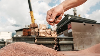 Detailansicht einer Hand die kleine Kupferkörner auf Haufen streut, im Hintergrund unscharf Industrieanlage mit gestapelten Kupfergegenständen