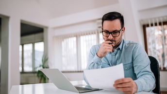 Person mit Brillen sitzt an Tisch, hält eine Hand mit Stift an Kinn und blickt auf Dokument, nebenstehend aufgeklapptes Notebook