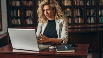 Person mit langen blonden Haaren sitzt an einem Schreibtisch, spricht und blickt dabei auf einem Laptop, während sie Kopfhörer trägt, ein Notizbuch, Stift und Smartphone liegen ebenfalls am Tisch, im Hintergrund zeigt sich ein Bücherregal