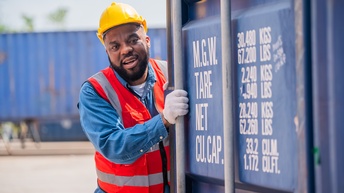 Eine Person in Arbeitskleidung mit Helm steht neben einem Container in einem Containerlager. Die Person berührt mit beiden Händen den Container.