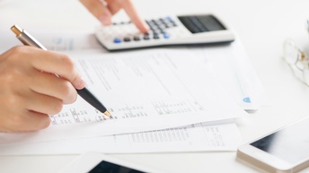 Nahaufnahme von Händen einer Person, in der rechten Hand hält sie einen Kugelschreiber, mit dem sie ein Formular ausfüllt. Mit der linken Hand tippt sie in einen Taschenrechner, der auf einem weißen Untergrund liegt. Rechts im Bild ist ein Smartphone